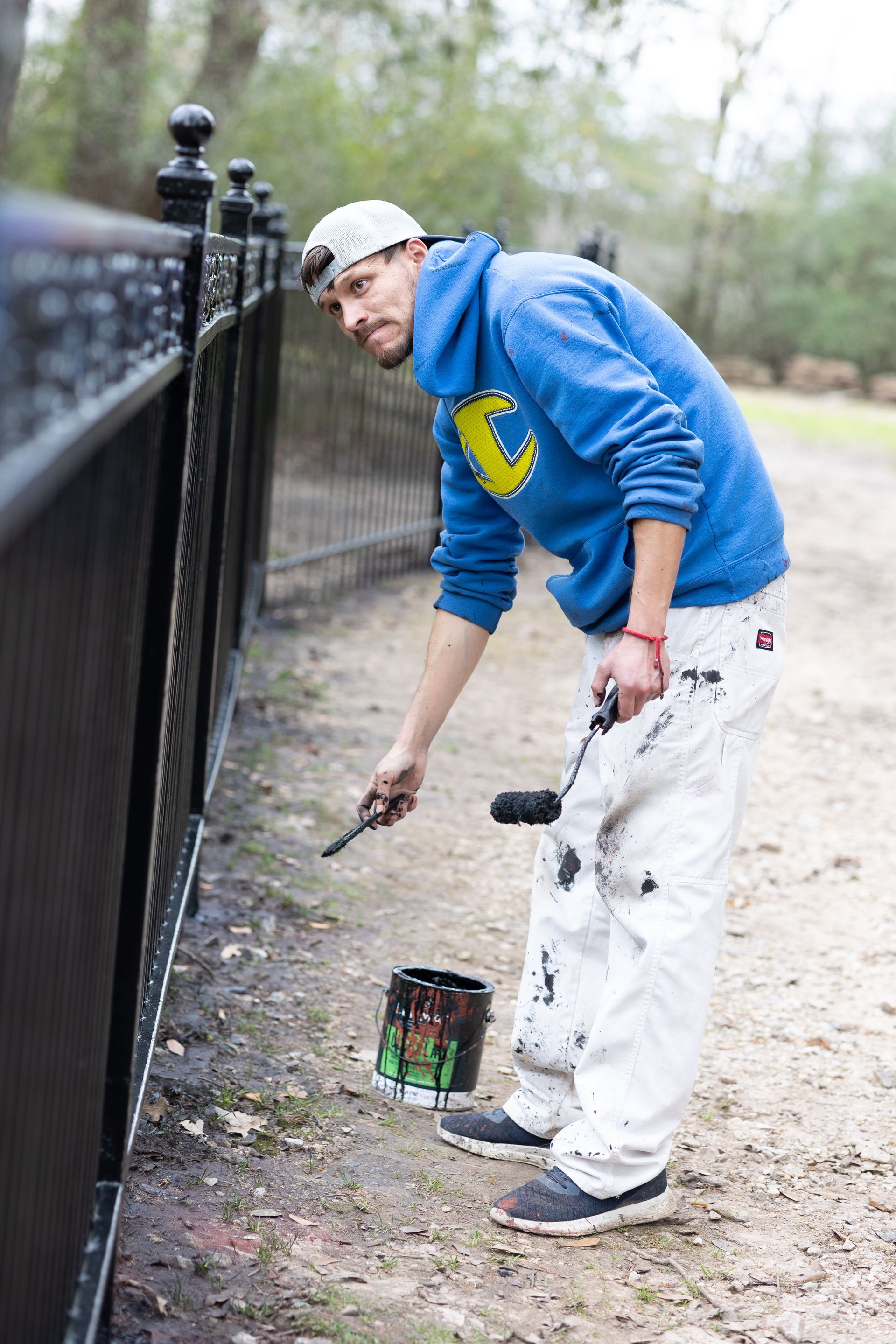 A man in a blue hoodie is painting a fence with a brush.