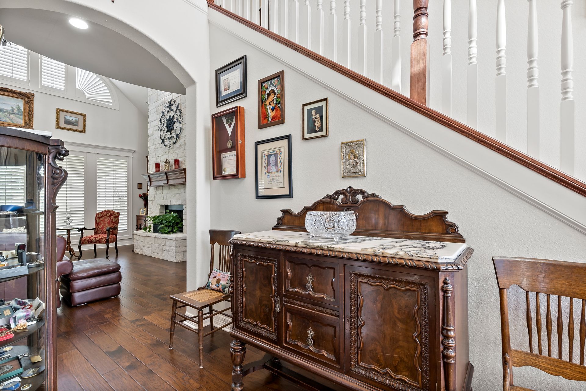 A living room with a staircase , a dresser , chairs and a clock on the wall.