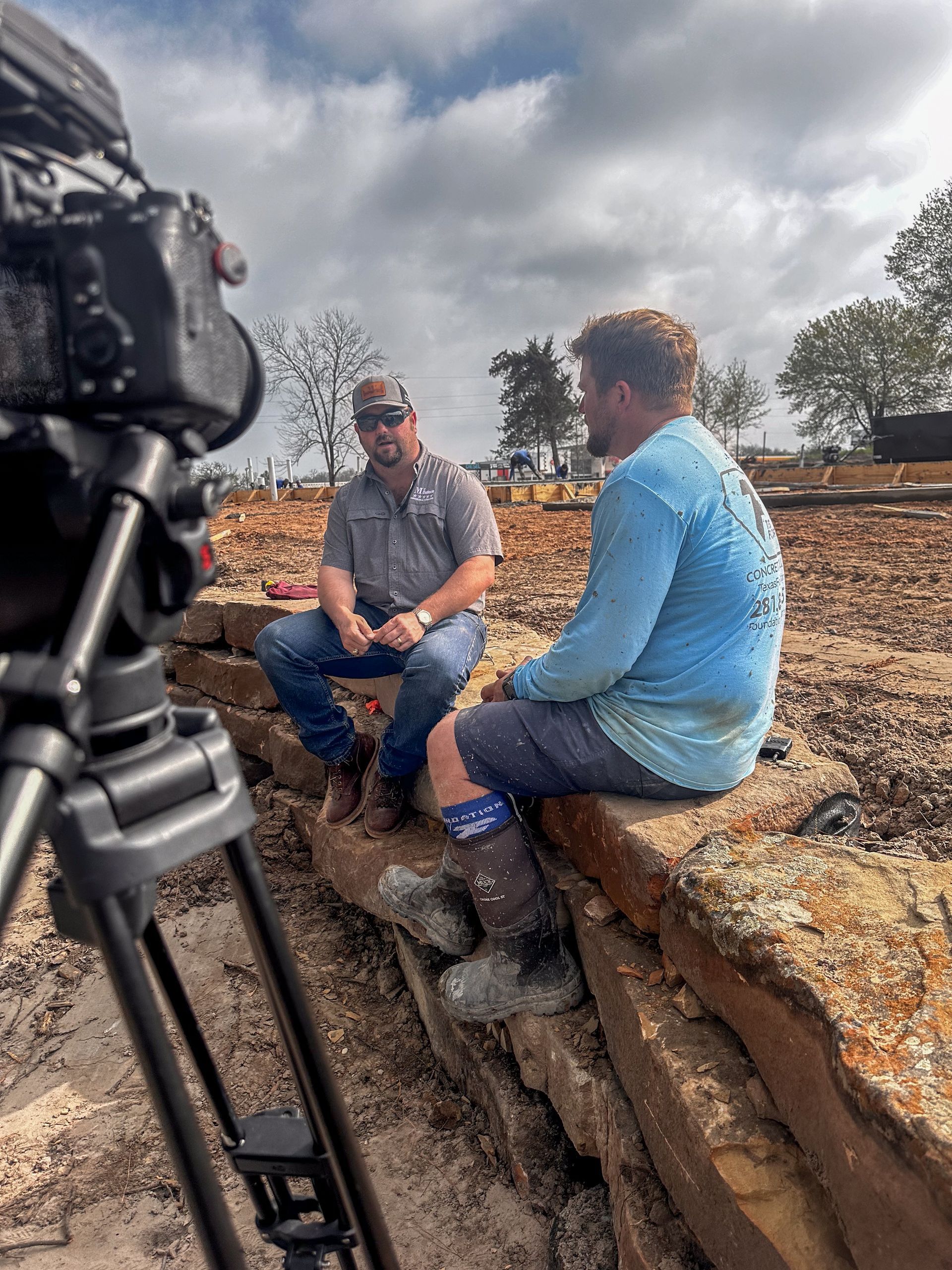 Two men are sitting on a rock talking to each other in front of a camera.
