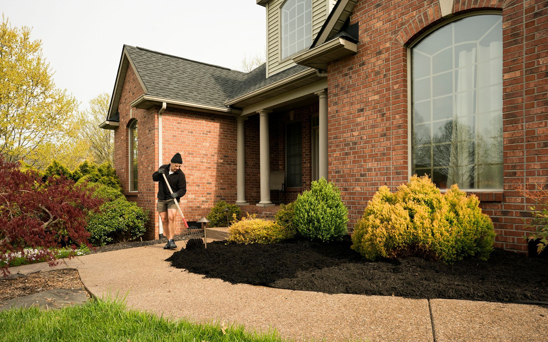 A man is raking mulch in front of a brick house.