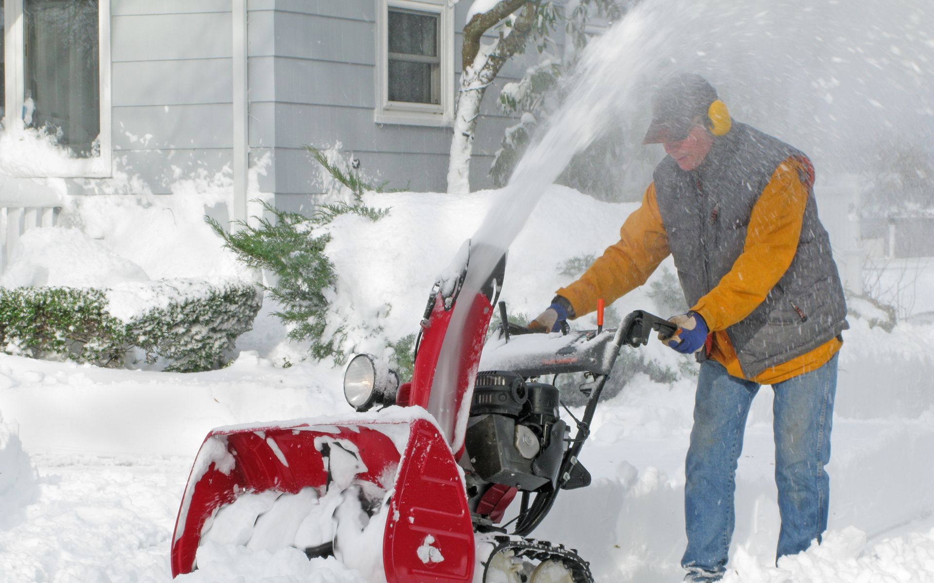 A man is blowing snow from a snow blower in front of a house.