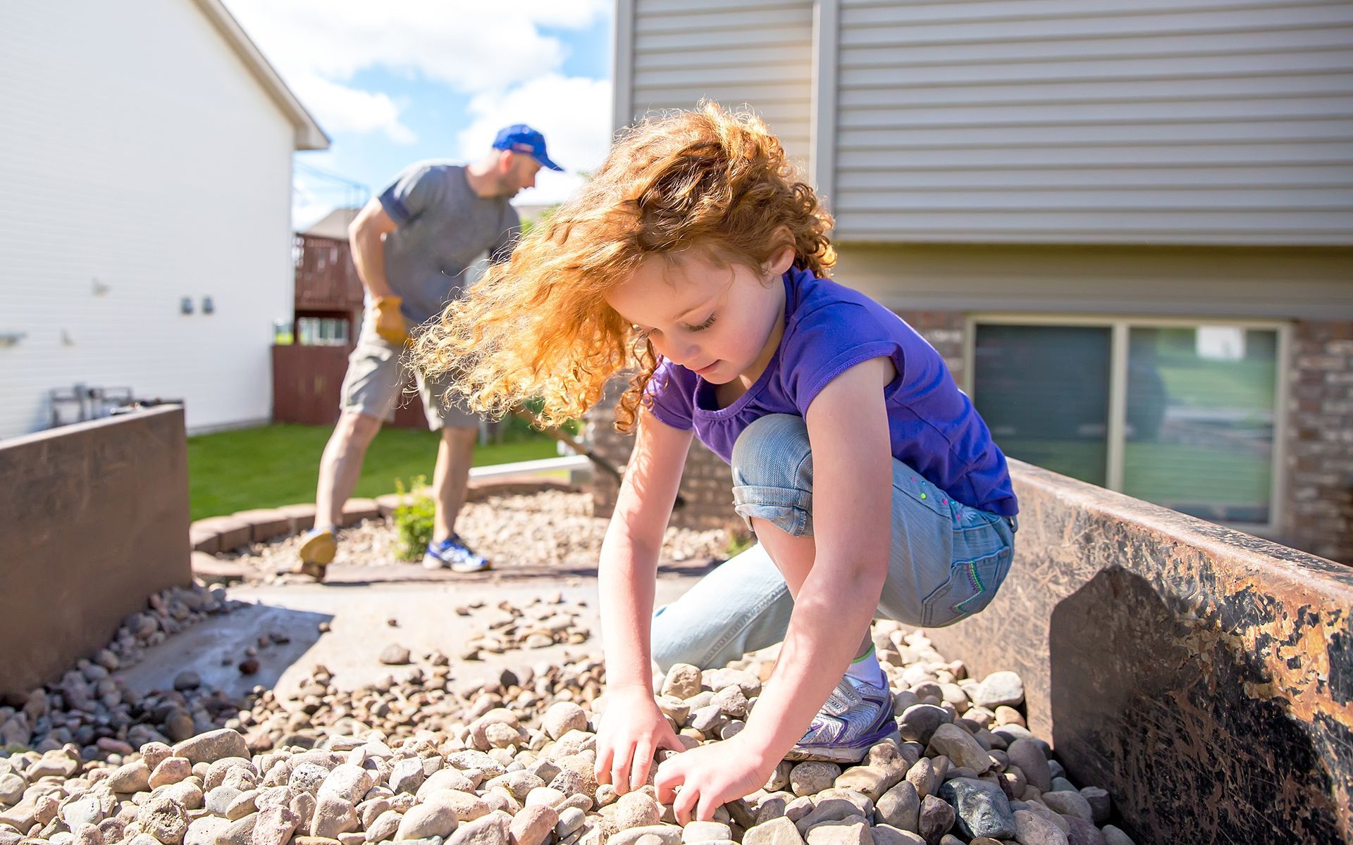 A little girl is playing with rocks in a yard while a man looks on.