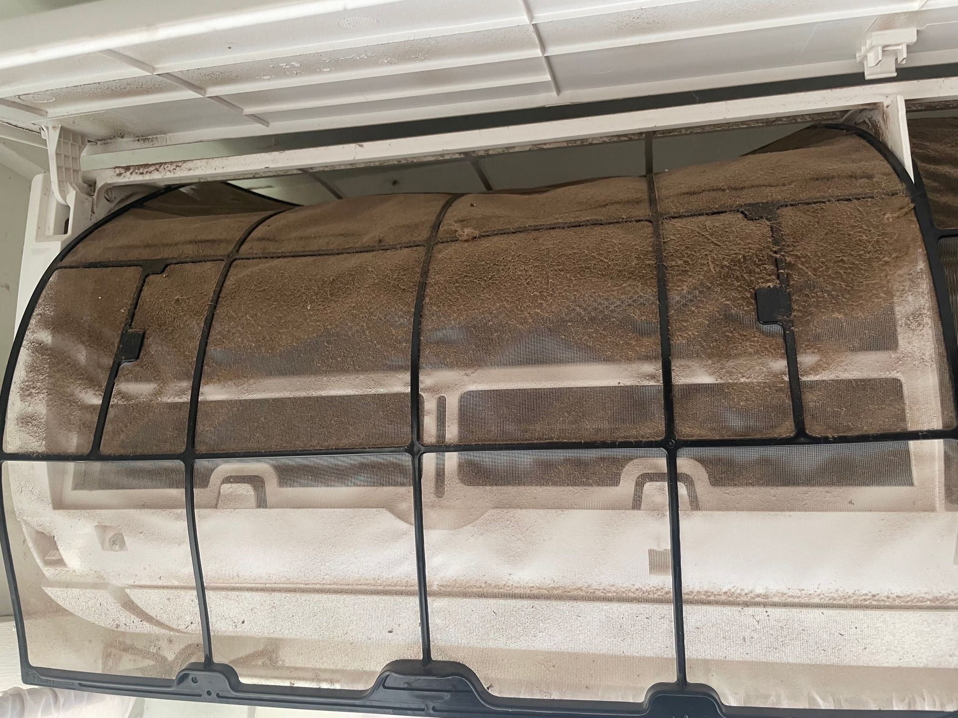 A heavily soiled air conditioner filter, covered in dust and debris, inside its unit.