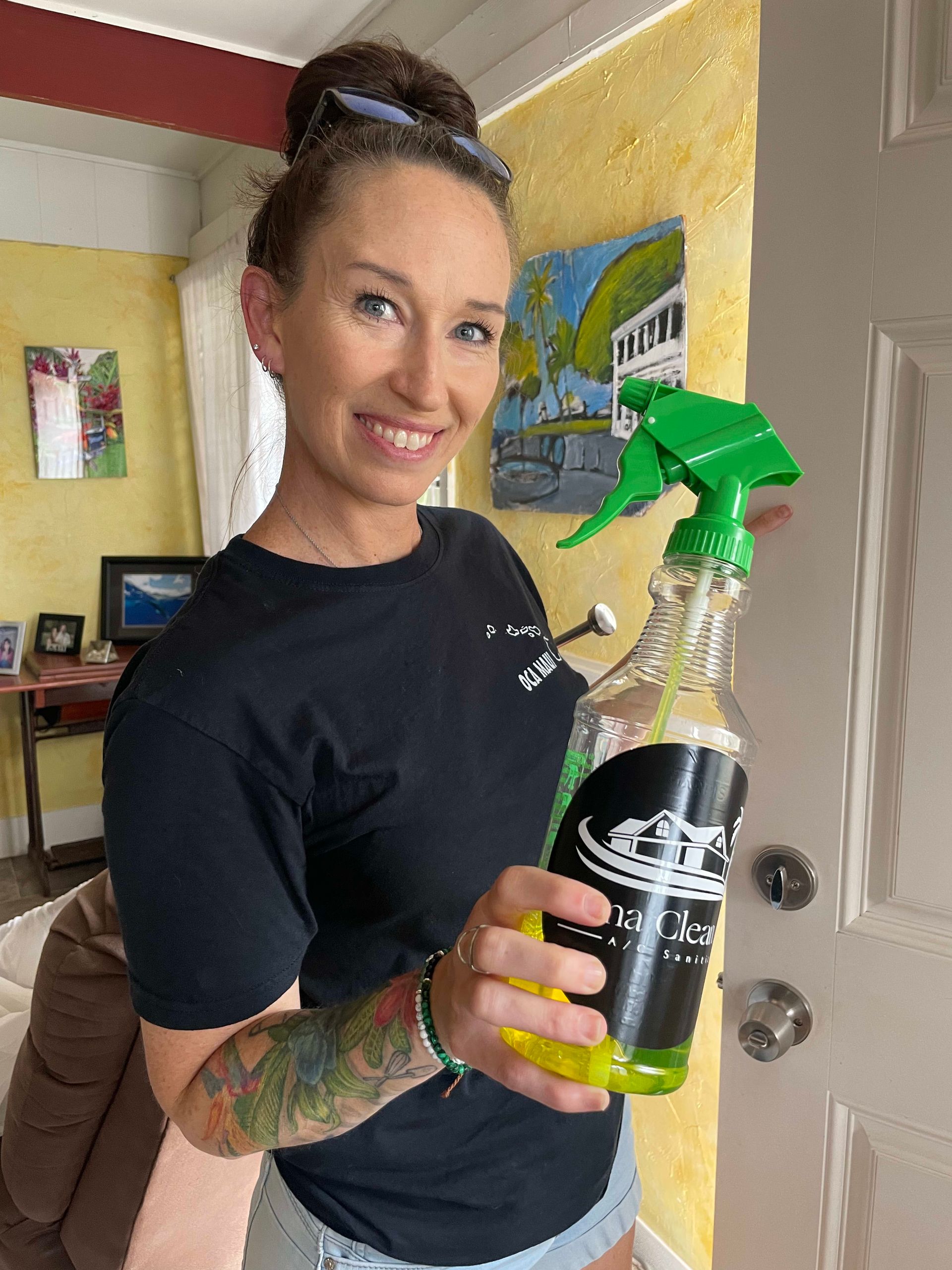 Woman smiling, holding a spray bottle in a brightly lit room. She wears a black shirt, has a tattoo, and hair up in a bun.