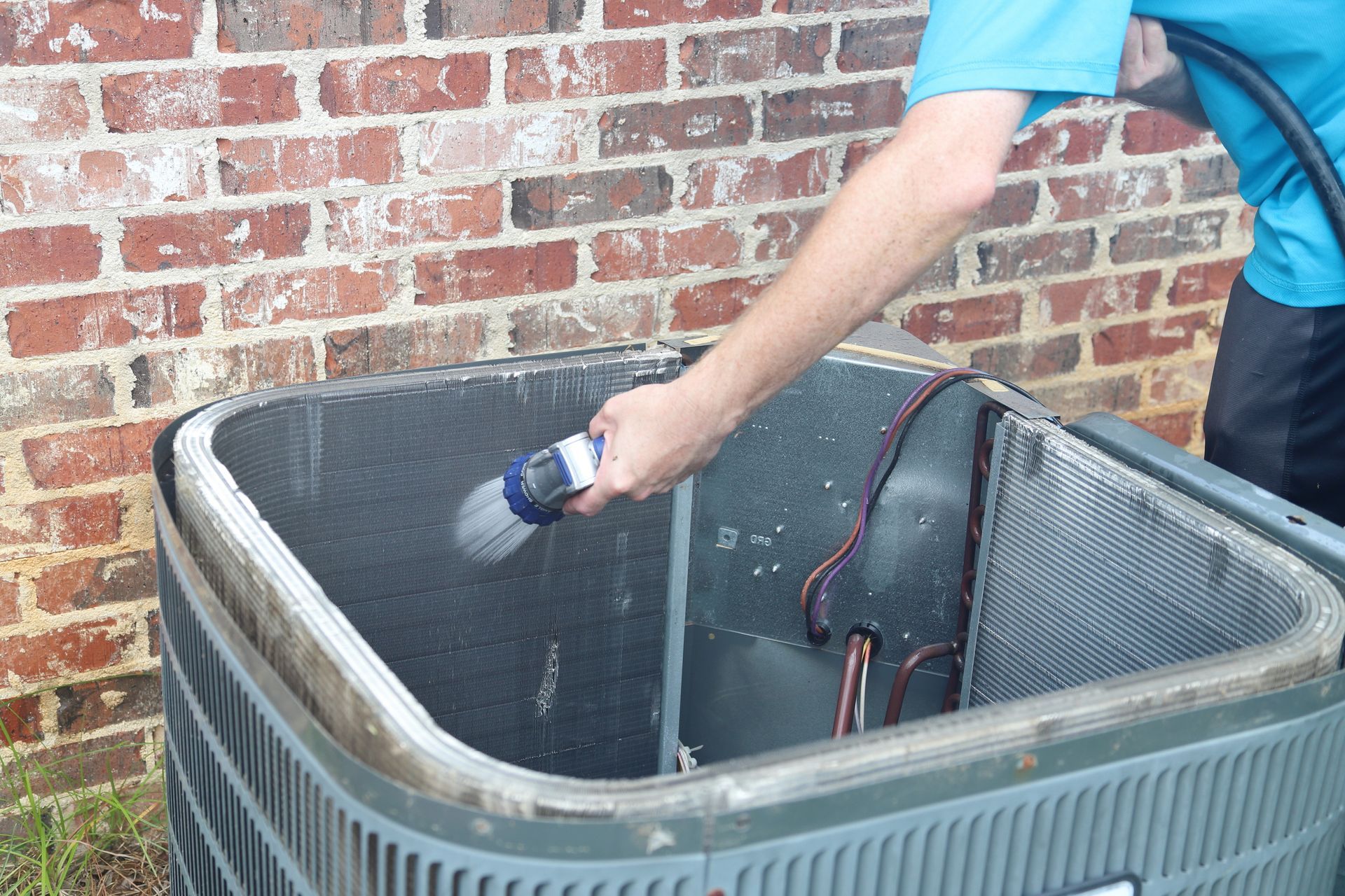 Person cleaning an air conditioning unit with a hose near a brick wall.
