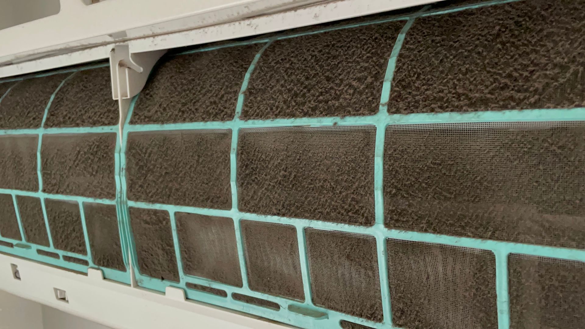 Close-up of a dirty air conditioner filter, covered in gray dust, in a white unit.