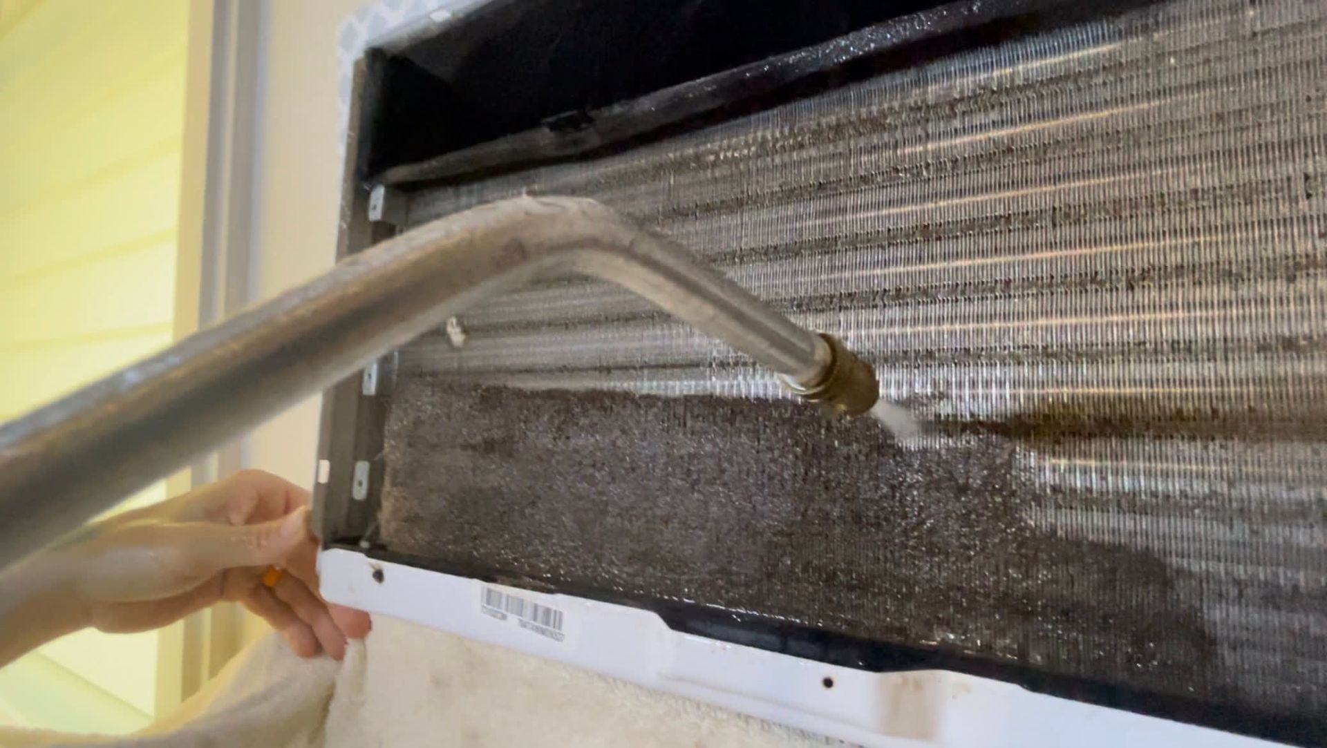 Person cleaning a dirty air conditioner with a nozzle, water spraying onto the coils.