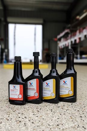Four Valvoline bottles of fuel additive. Black bottles with colored labels sit on a speckled floor, garage in background.