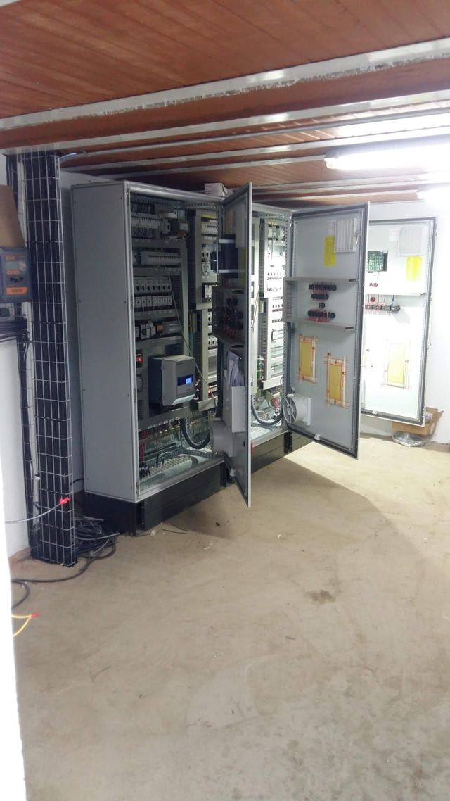 Paneles de control eléctrico abiertos en un cuarto de servicio con cableado expuesto y piso de concreto.