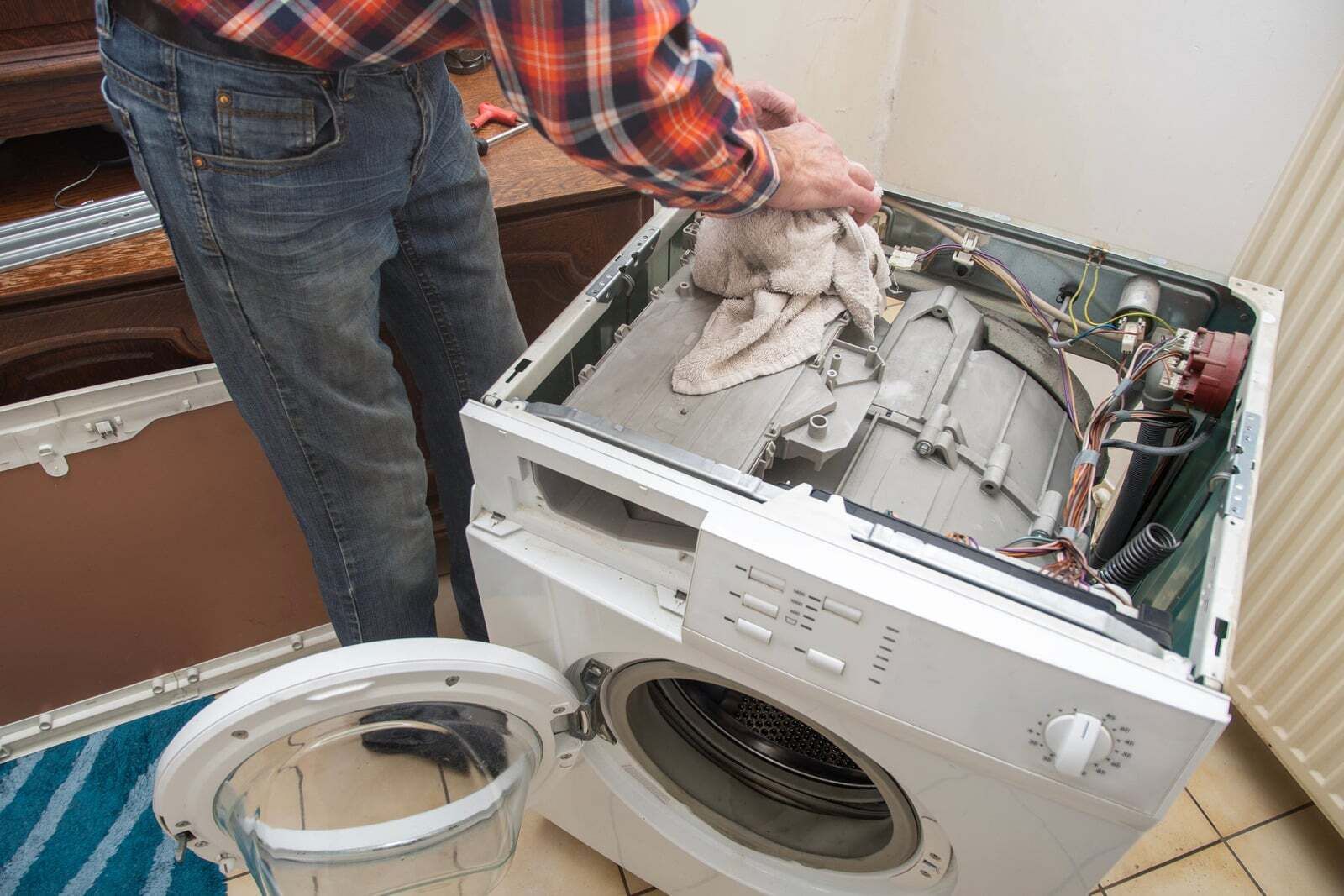 Person repairing a white dryer with its top open, exposing the drum and internal parts.