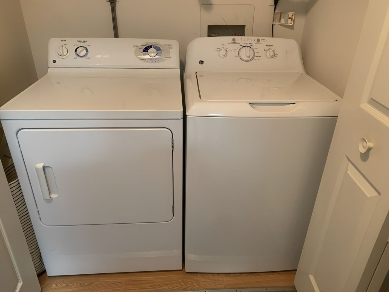Laundry room with two white front-loading machines, white tiled walls, dark floor, and a small plant on top.