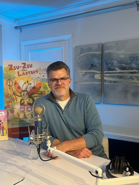 Man at a desk with a microphone, book poster behind him. Indoor setting with blue lighting.
