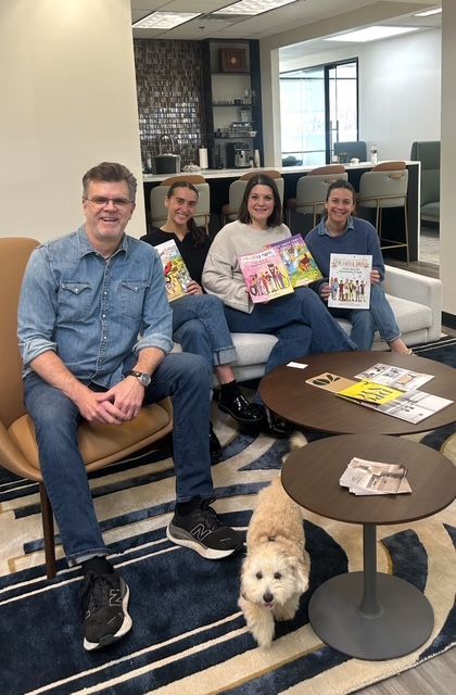 Four people and a dog in an office, holding books and magazines. They are sitting on chairs and a sofa.