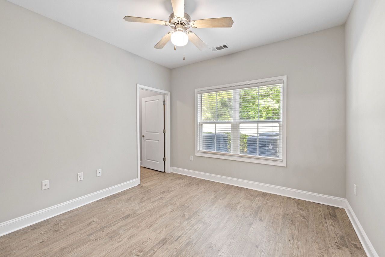 Empty bedroom with ceiling fan, large window, and light wood flooring.
