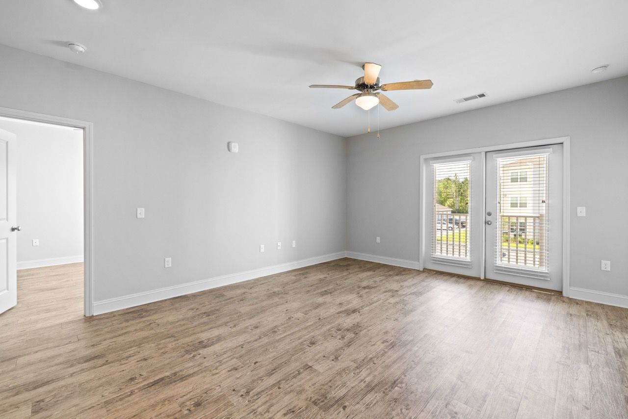 Empty living room with light gray walls, wood-look flooring, ceiling fan, and balcony doors.