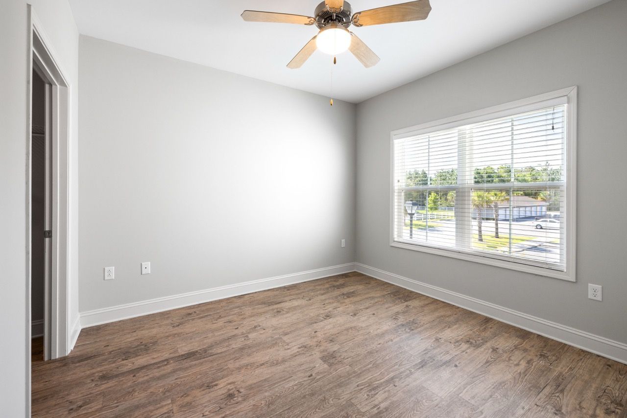 Empty bedroom with a large window, light gray walls, wood flooring, and a ceiling fan.