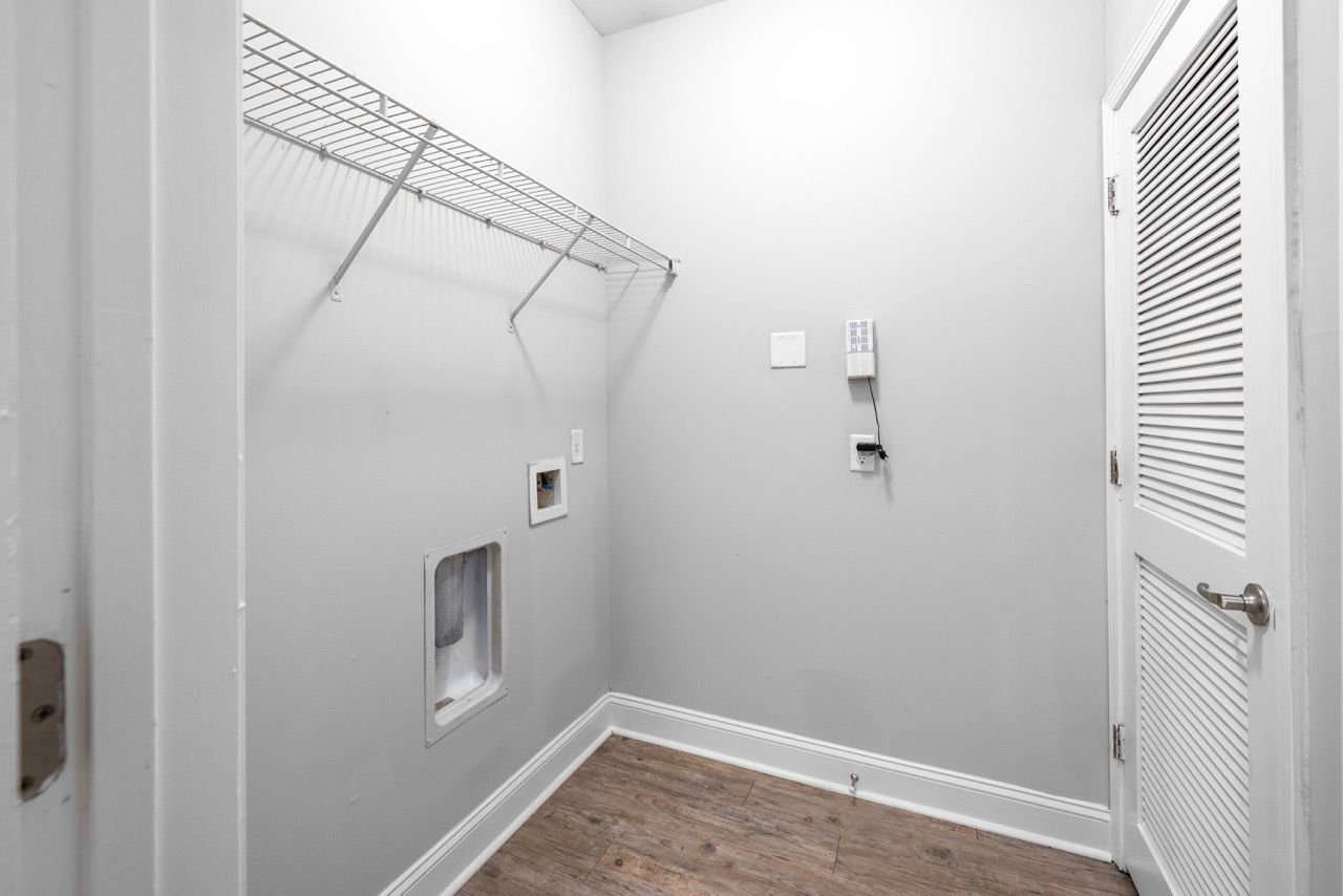 Laundry closet with wire shelf and washer/dryer hookups, white walls, and a louvered door.