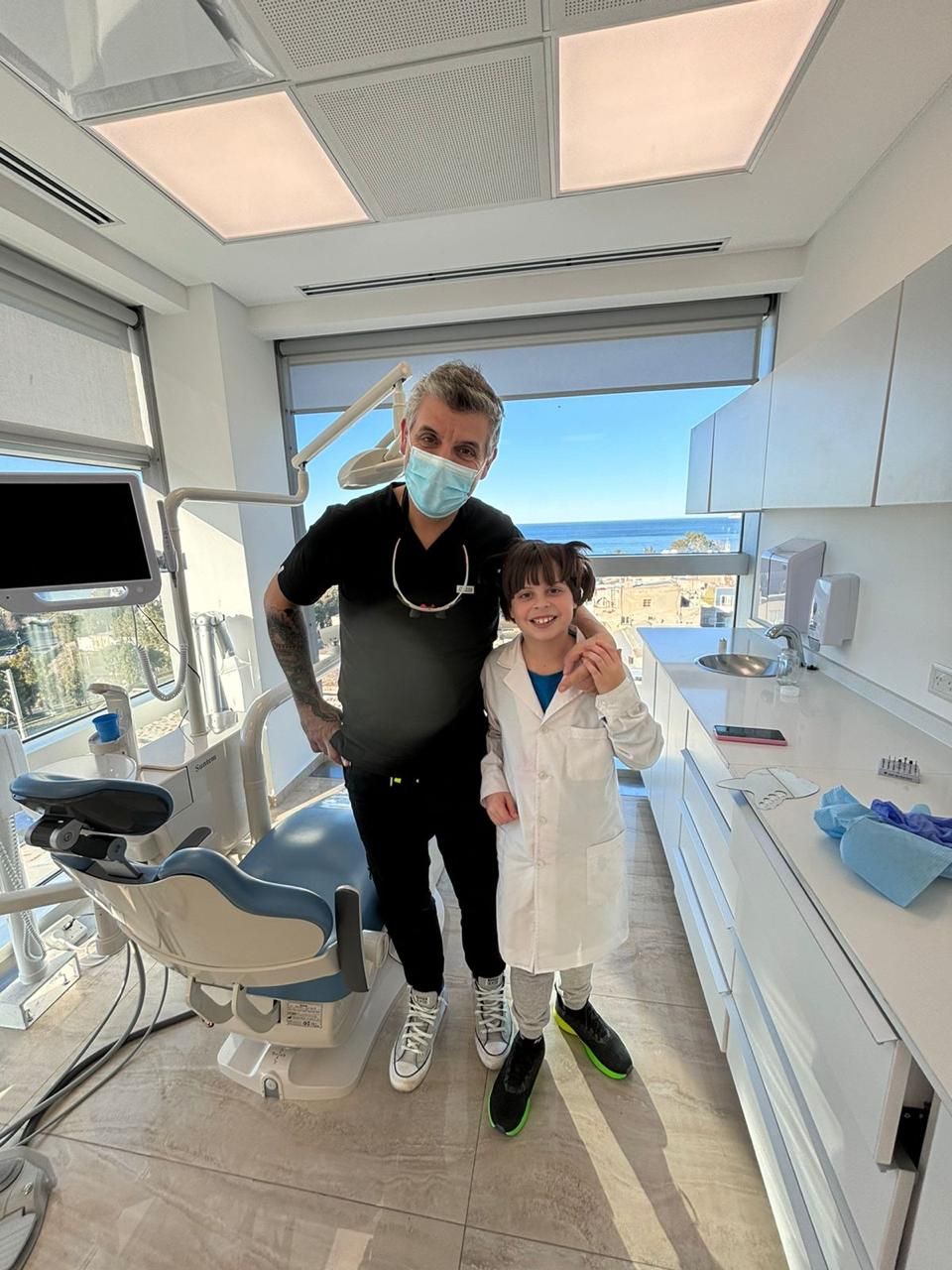 Dentista y niño con bata de laboratorio posan en un consultorio dental moderno con vista al mar.
