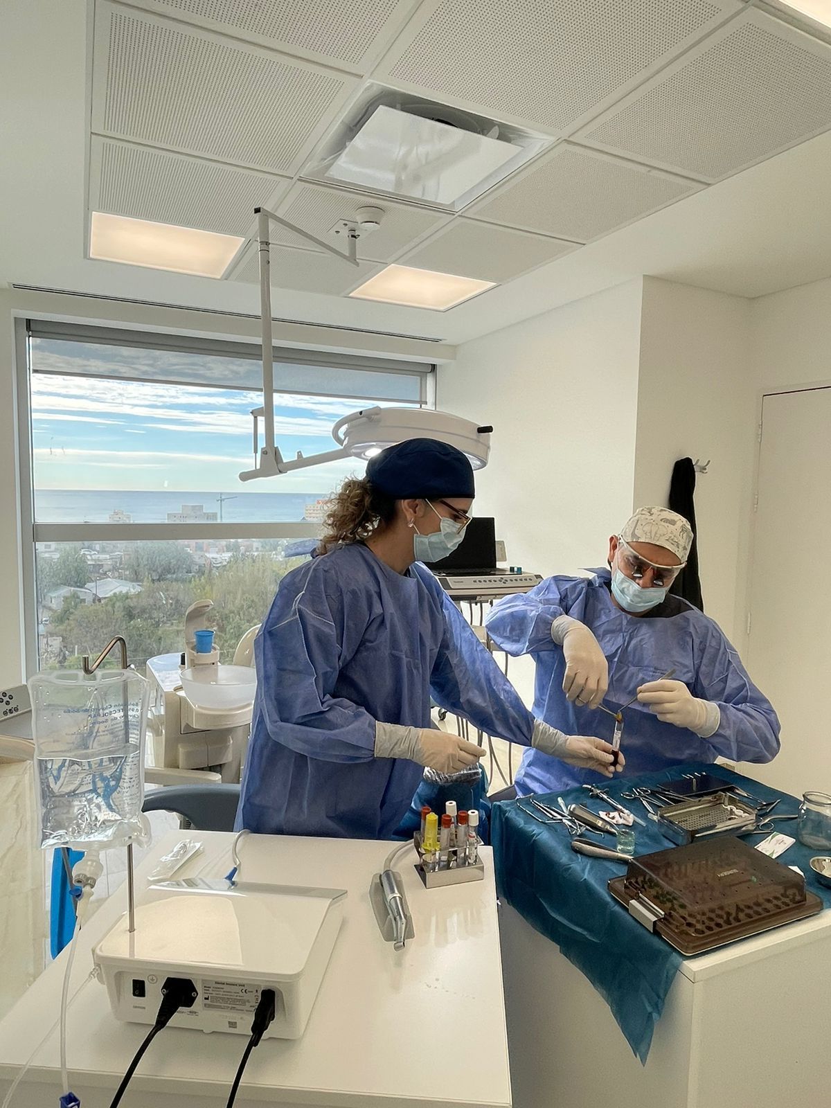 Dos cirujanos vestidos de quirofano preparan instrumentos en un quirófano, con una ventana que da a un paisaje urbano.