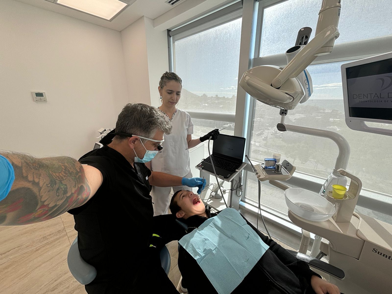 Dentista tomándose una selfi durante una intervención dental. Asistente presente. Consultorio dental luminoso.