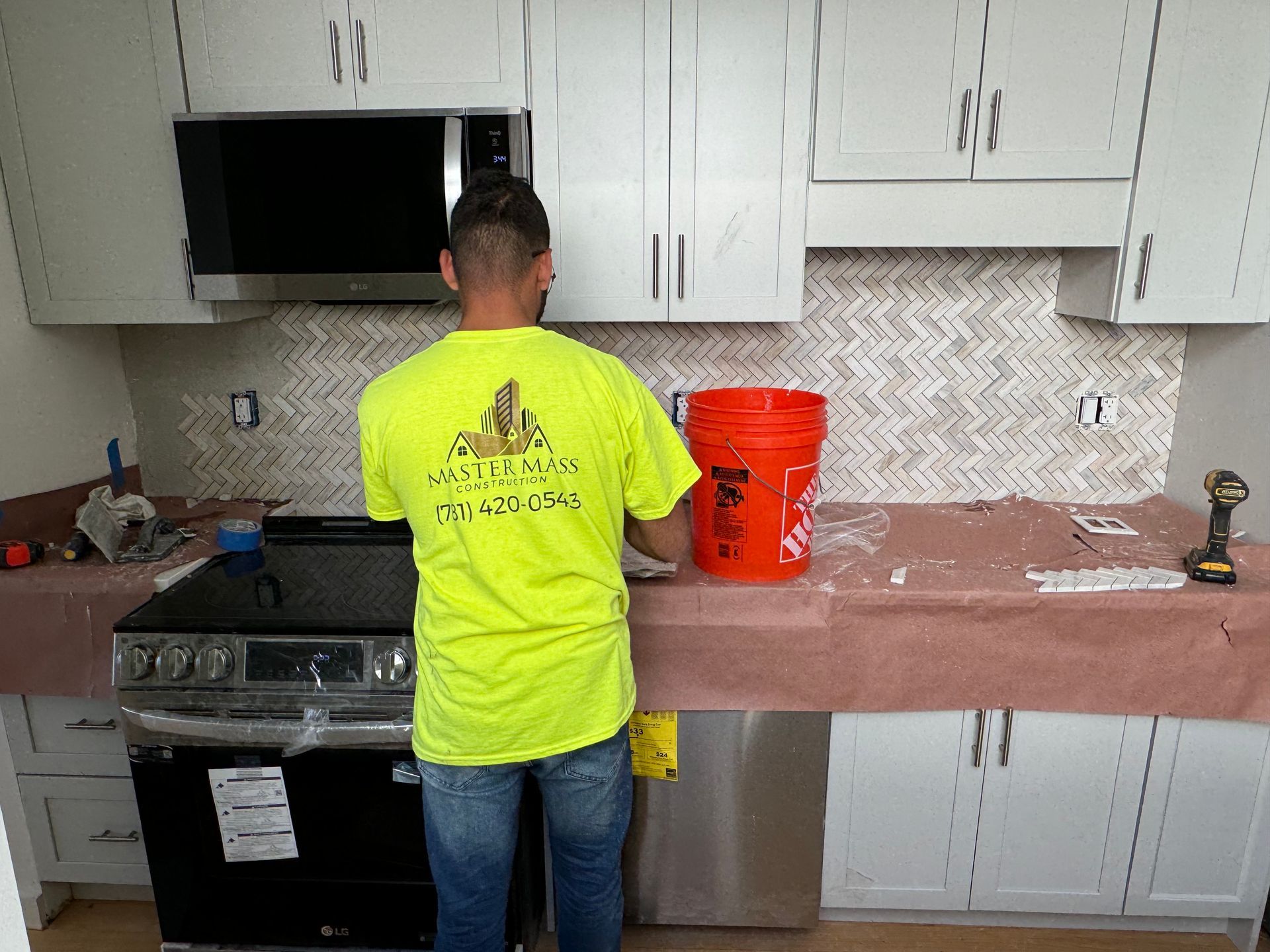 Man tiling a kitchen backsplash, standing in front of cabinets and a stove, holding a trowel next to a bucket.