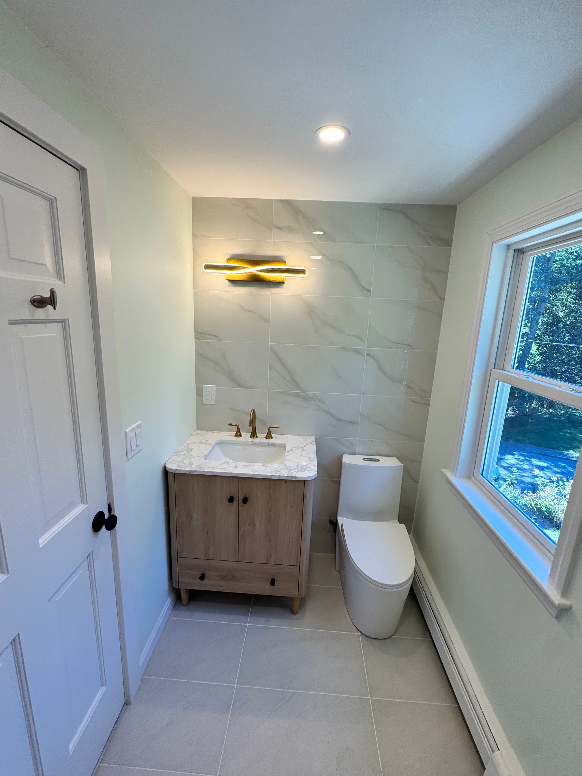 Bathroom with light green walls, wood vanity, marble-like tiled wall, modern toilet, and small window.
