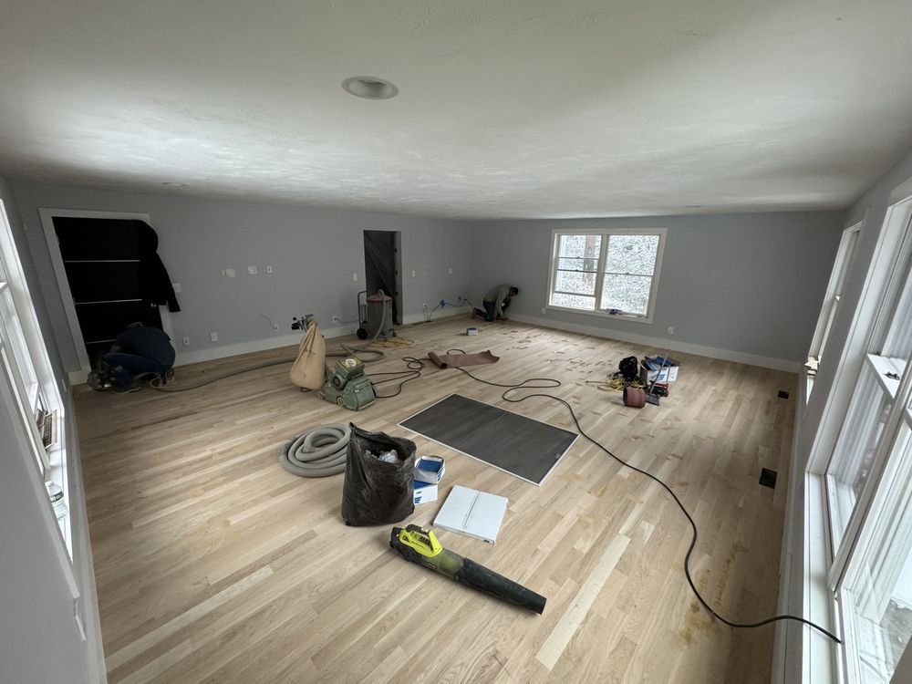 Living room renovation in progress, light wood flooring, gray walls, tools and materials scattered.