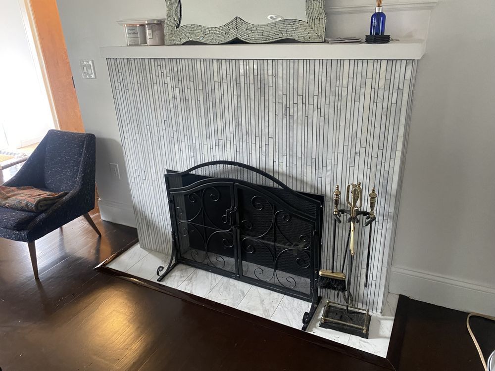 Fireplace with a black screen and tools; white and gray textured facade; blue chair in the corner.