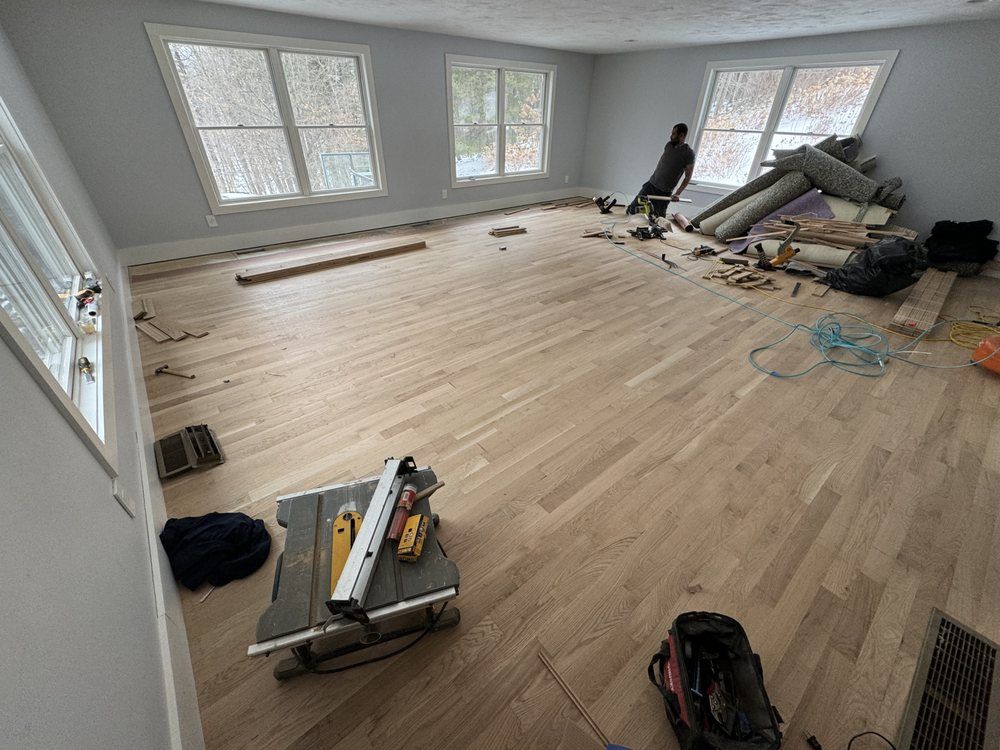 Room with light wood floor, windows, and construction tools. A person sits near a pile of materials.