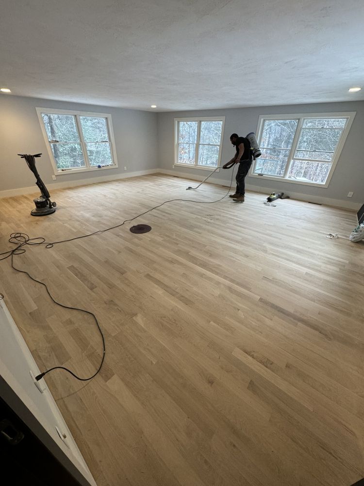Person applying finish to newly sanded hardwood floor in a room with windows.