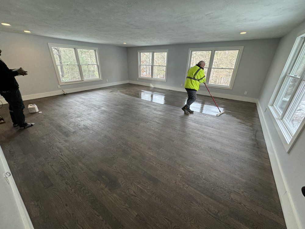 Two people applying sealant to a dark wood floor in a room with several windows.