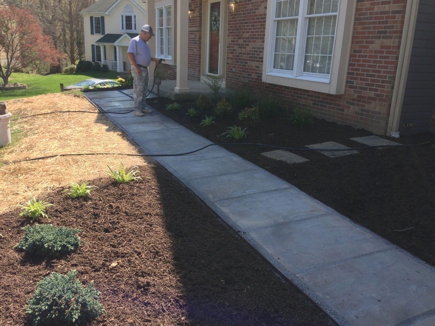 Man waters a newly landscaped walkway and flowerbeds in front of a brick house.