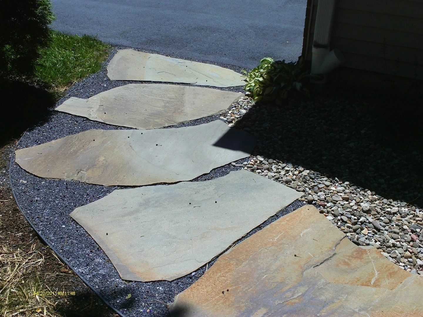 Stone path through dark gray gravel, leading to a house, with green grass on the left.