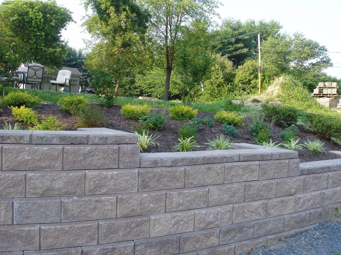 Stone retaining wall with a landscaped hillside featuring bushes and a tree.