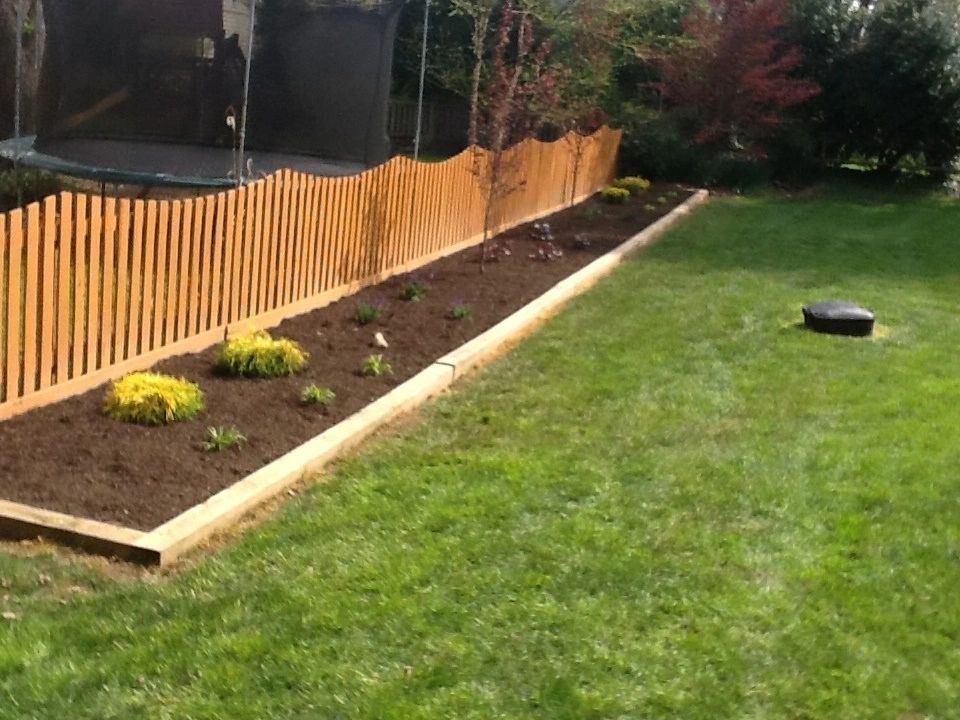 Wooden fence and flower bed along green lawn, trampoline in background.