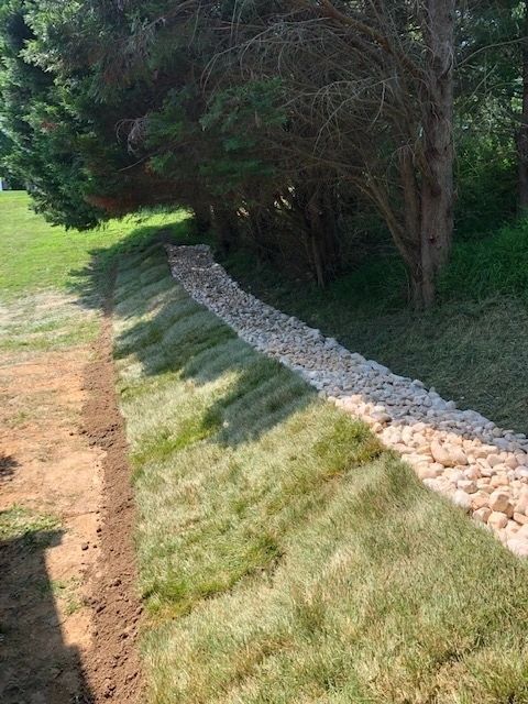 Grassy hill with rock-lined channel under trees; freshly planted grass on the slope.