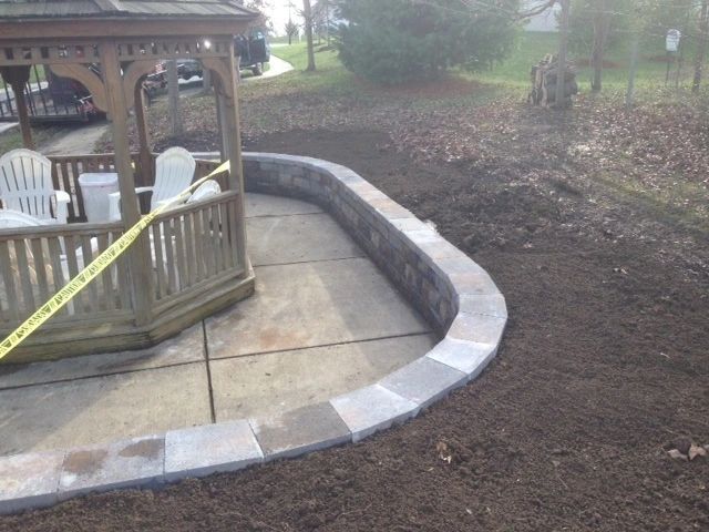 Stone retaining wall curves around a concrete patio with a wooden gazebo and lawn.