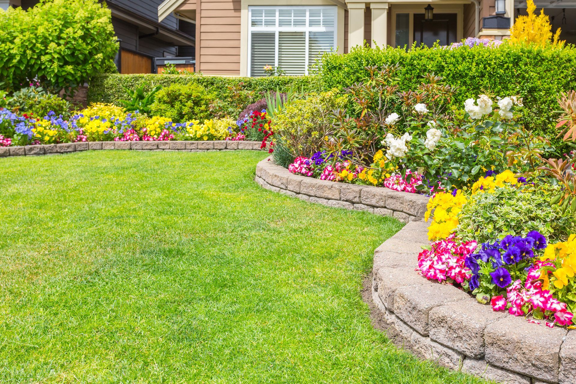 Lush green lawn with colorful flower beds edged by a stone border in front of a house.