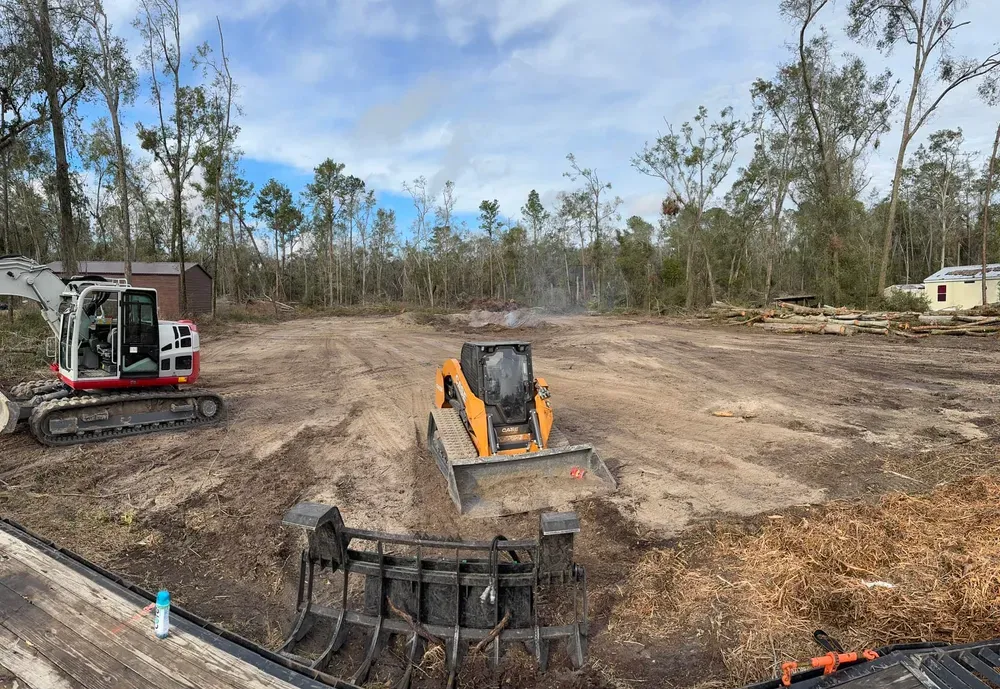 Construction site with two machines clearing land: excavator left, skid steer center, trees in the background.