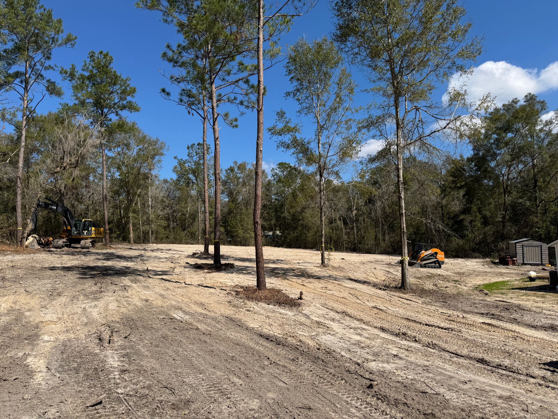 Construction site with dirt ground and scattered trees. Machinery is present. Blue sky.