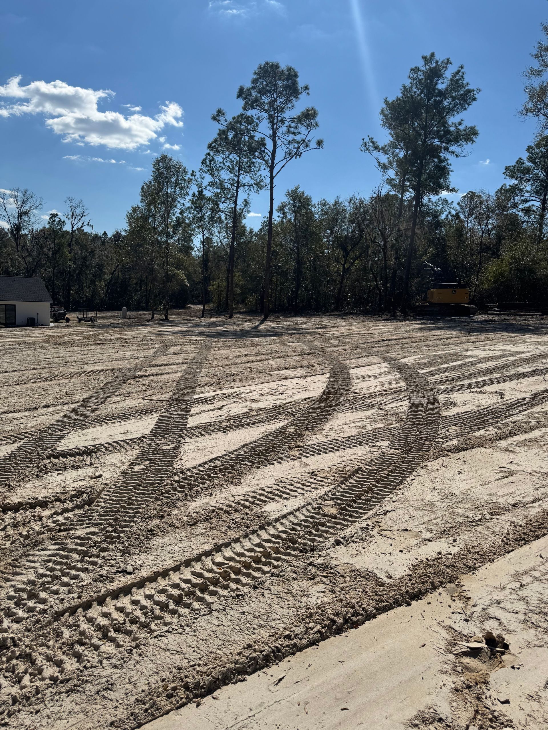 Sandy lot with tire tracks, trees in the background, and a partially visible structure on a sunny day.