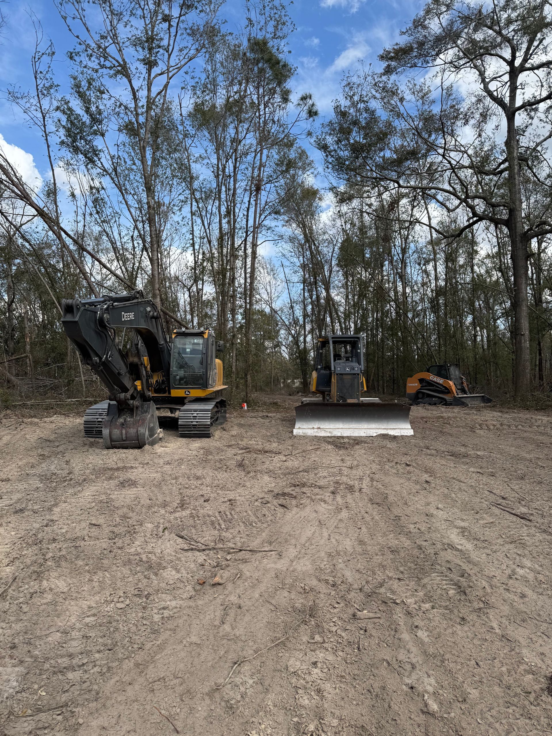 Construction site: Excavator and bulldozer on cleared land, trees in background under blue sky.