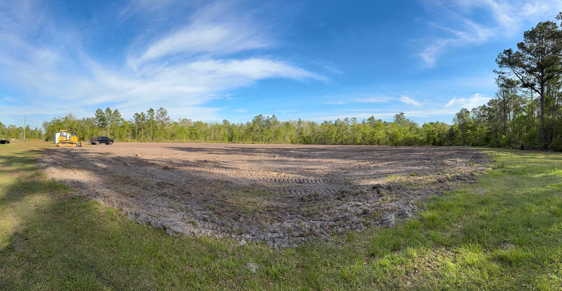 A large, cleared dirt area surrounded by grass and trees under a blue sky with clouds.