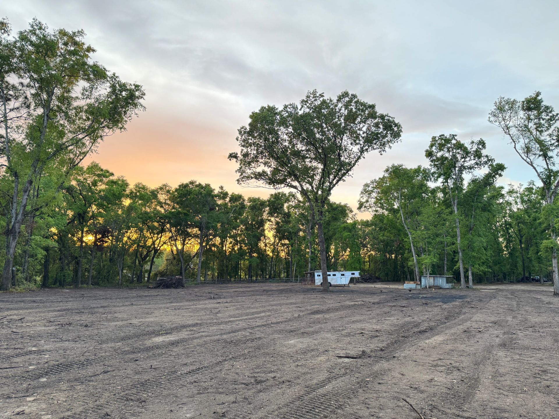 A sunset over a treeline, viewed from a leveled dirt ground, with some small buildings.