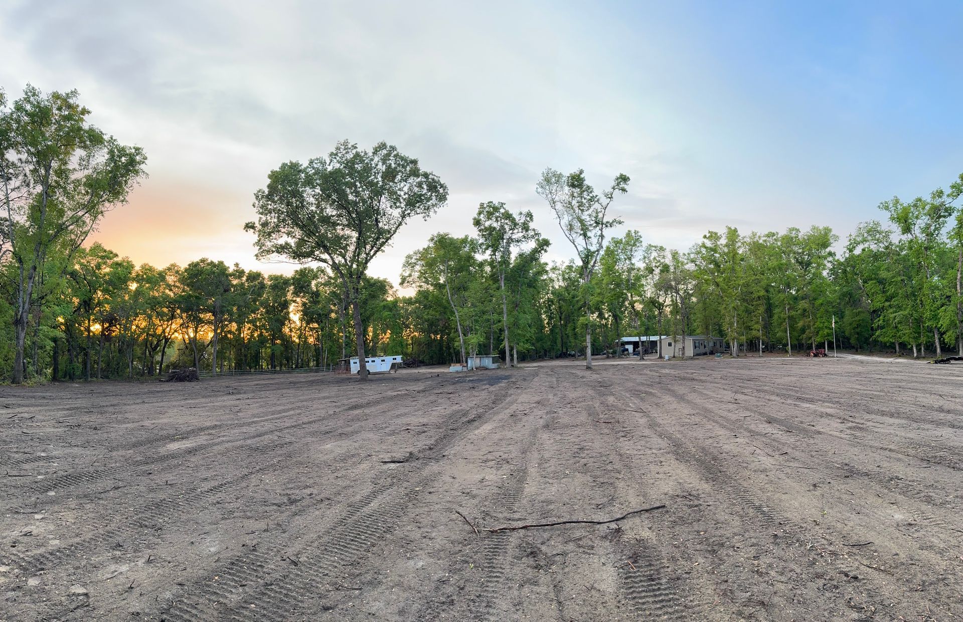 A cleared dirt lot at dusk with trees in the background and a light blue sky.