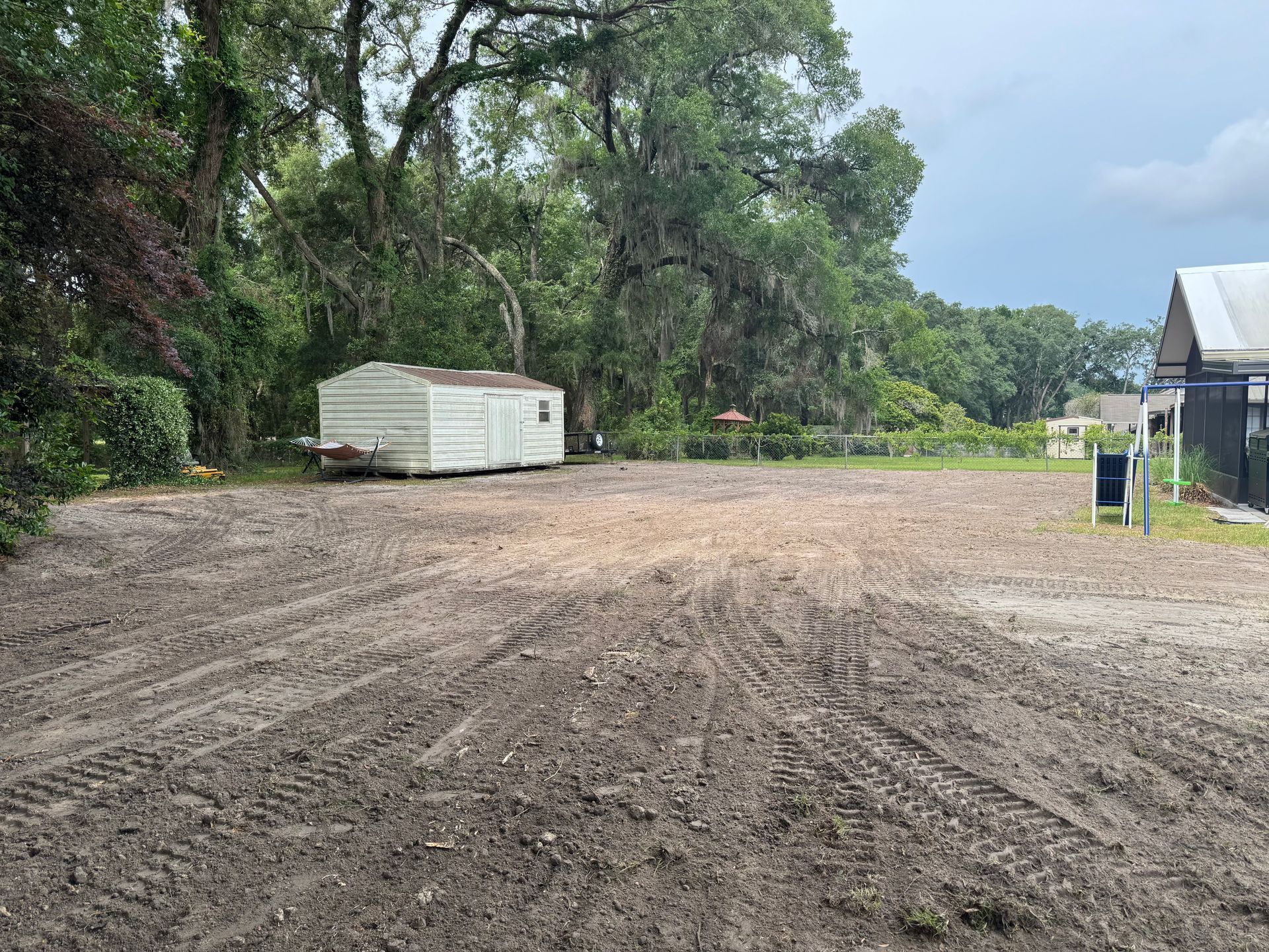 A cleared dirt lot with tire tracks, a small shed, and trees under a cloudy sky.