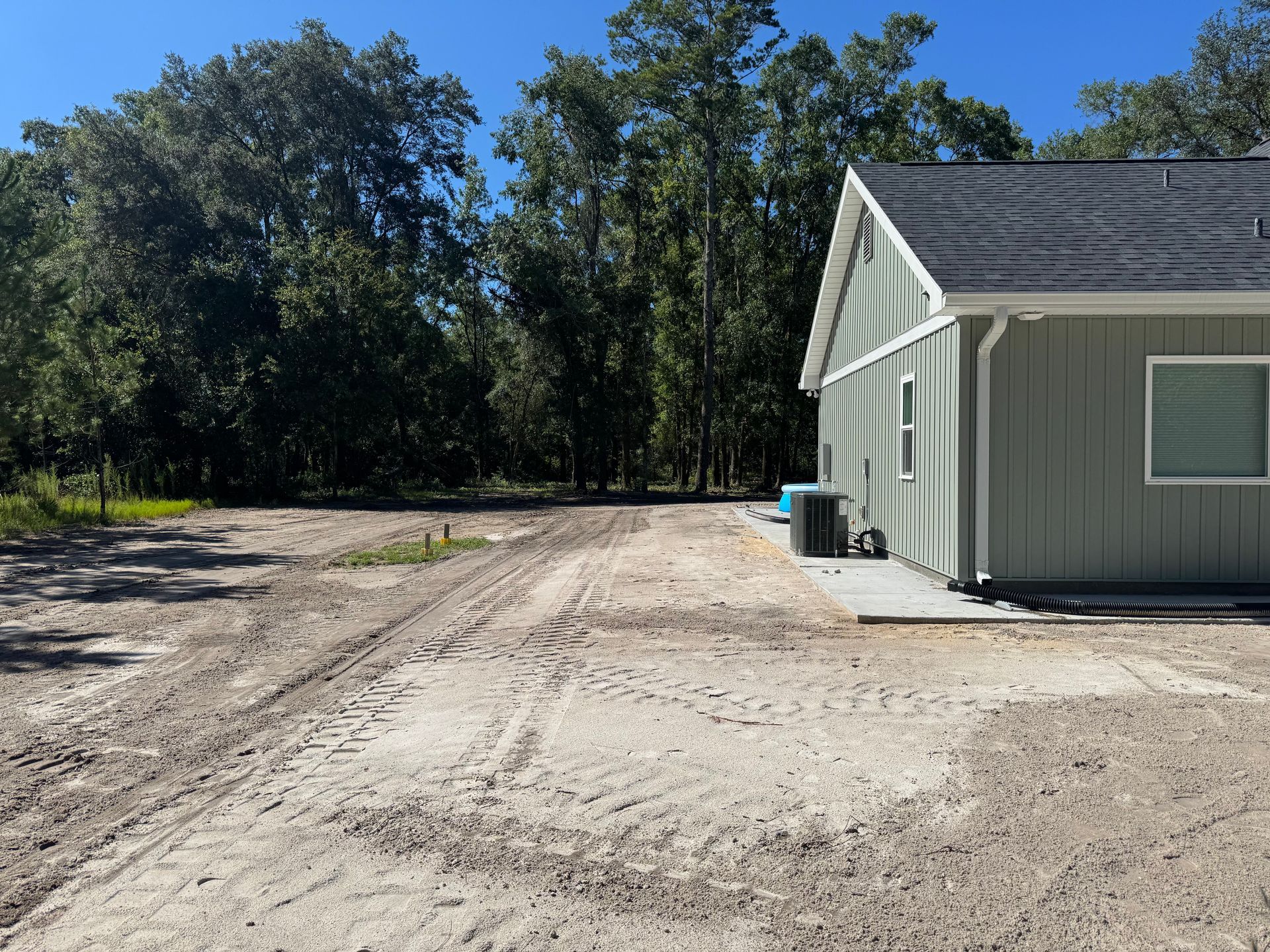 A dirt driveway next to a light green house, leading to a forest on a sunny day.