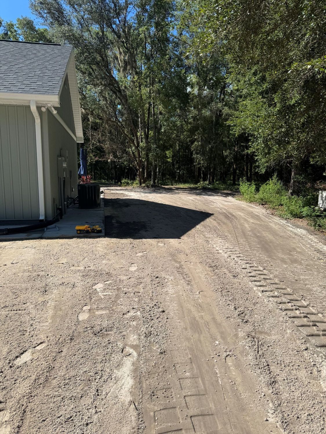 Gravel driveway beside a building with dark trash cans and leading into a wooded area, sunny day.