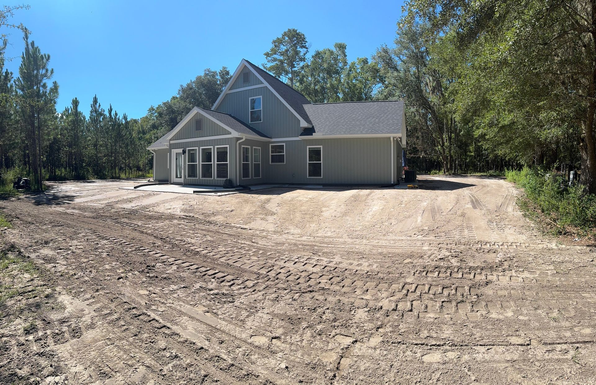 House under construction; dirt lot with tire tracks. Light green siding, gray roof, surrounded by trees.