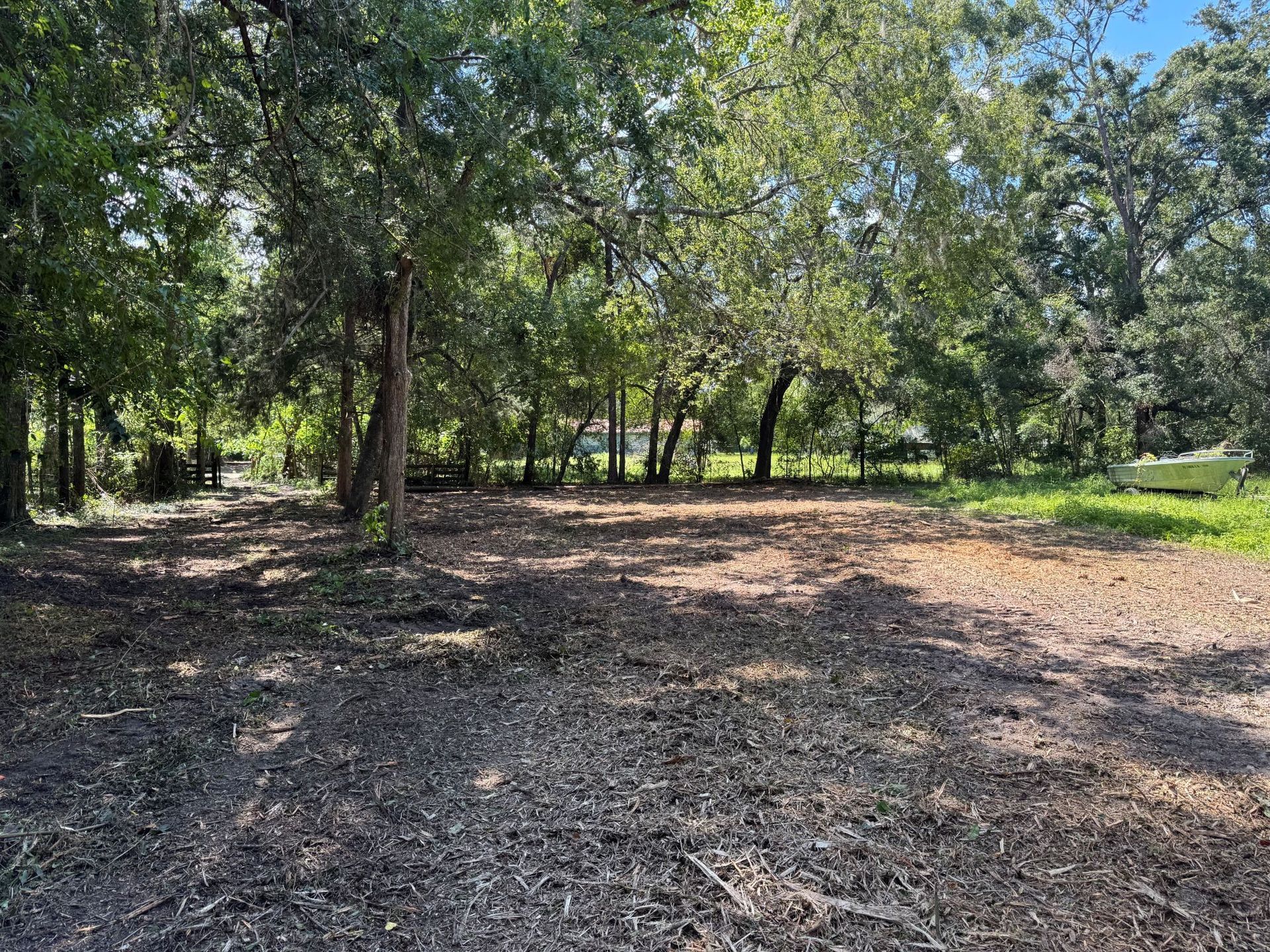 Cleared area in a wooded lot; brown mulch, green trees, and a glimpse of a boat.